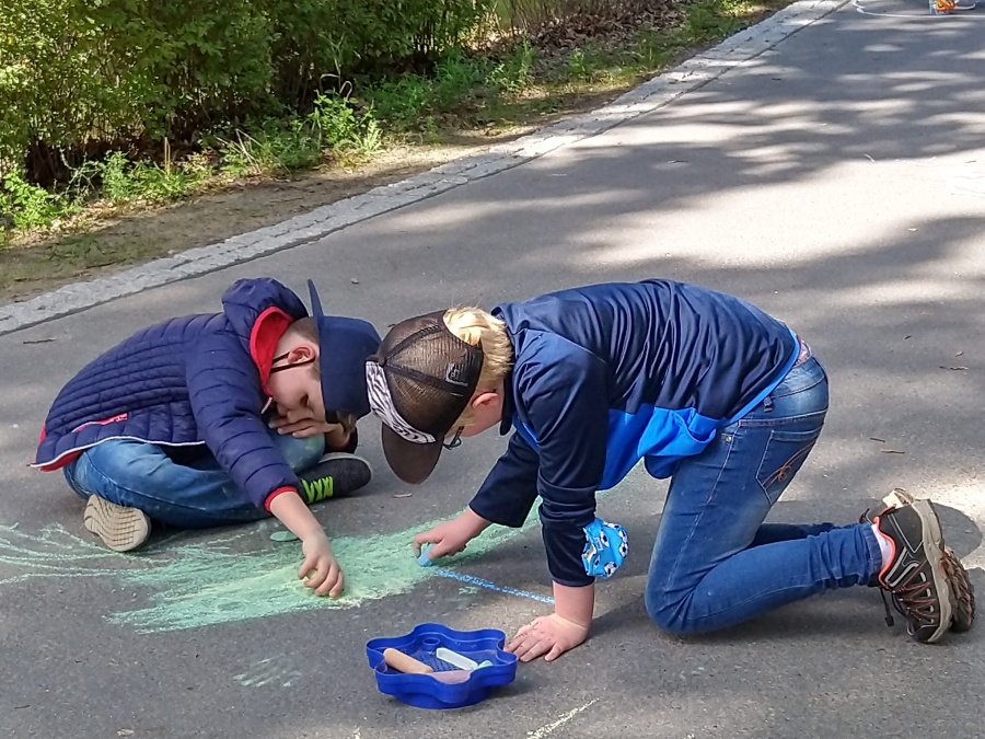 Zwei Kinder knien auf einem asphaltierten Weg und malen mit Straßenkreide Bilder.